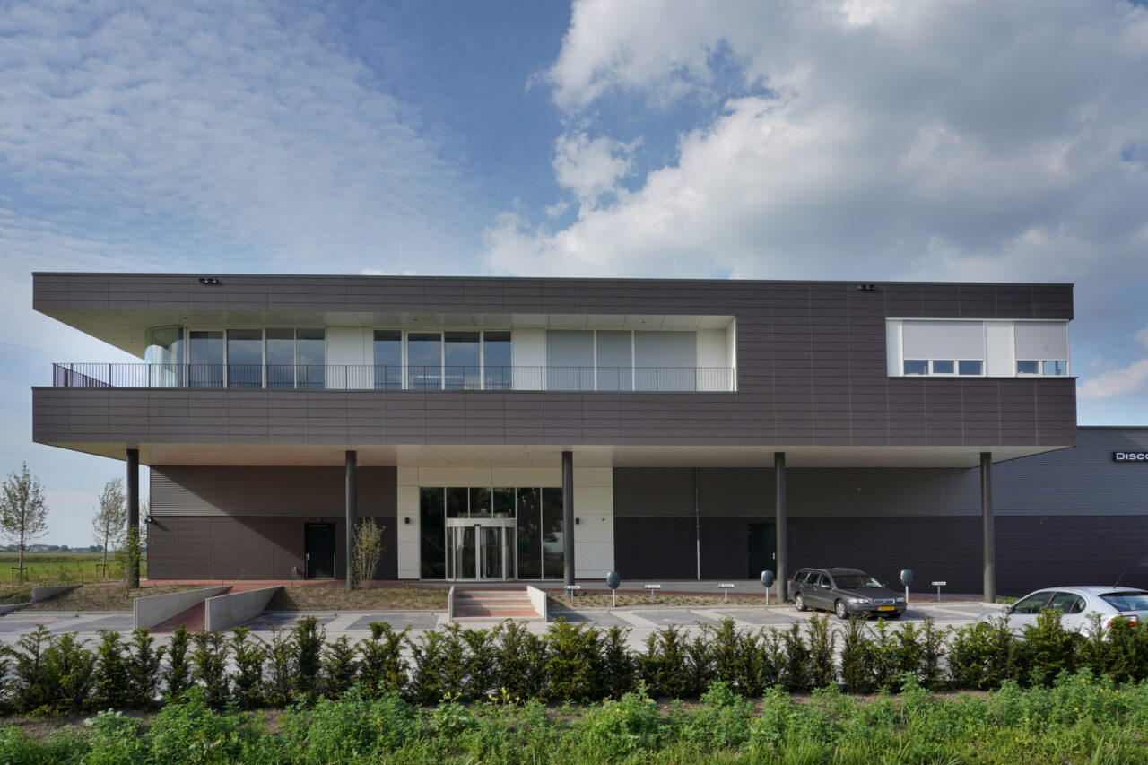 Frontansicht des Hapam-Gebäudes in Bunschoten-Spakenburg mit modernen dunklen Keramikpaneelen, großen Fenstern und einem überdachten Eingangsbereich, umgeben von einer gepflegten Grünfläche und Parkplätzen