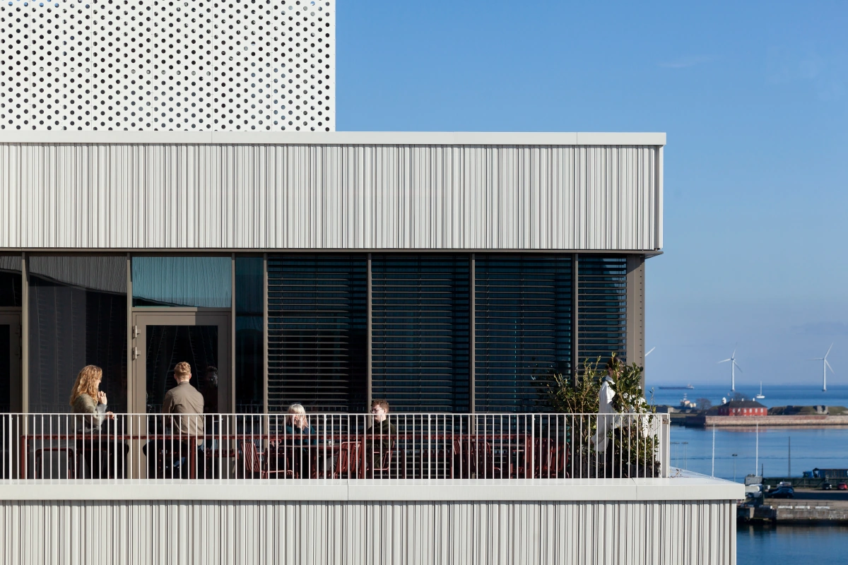 Gebäude mit weißer Keramikfassade und stark profiliertem Dach, vertikales Design bei Marmorvej, Menschen auf dem Balkon mit Blick auf das Meer und Windturbinen im Hintergrund